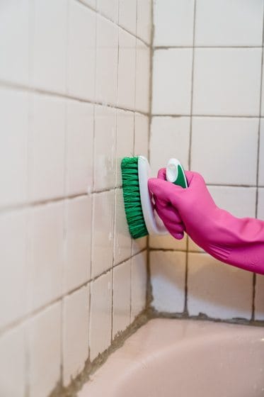 Gloved hand scrubbing mildew from shower tile grout in a Chicago bathroom with a cleaning brush